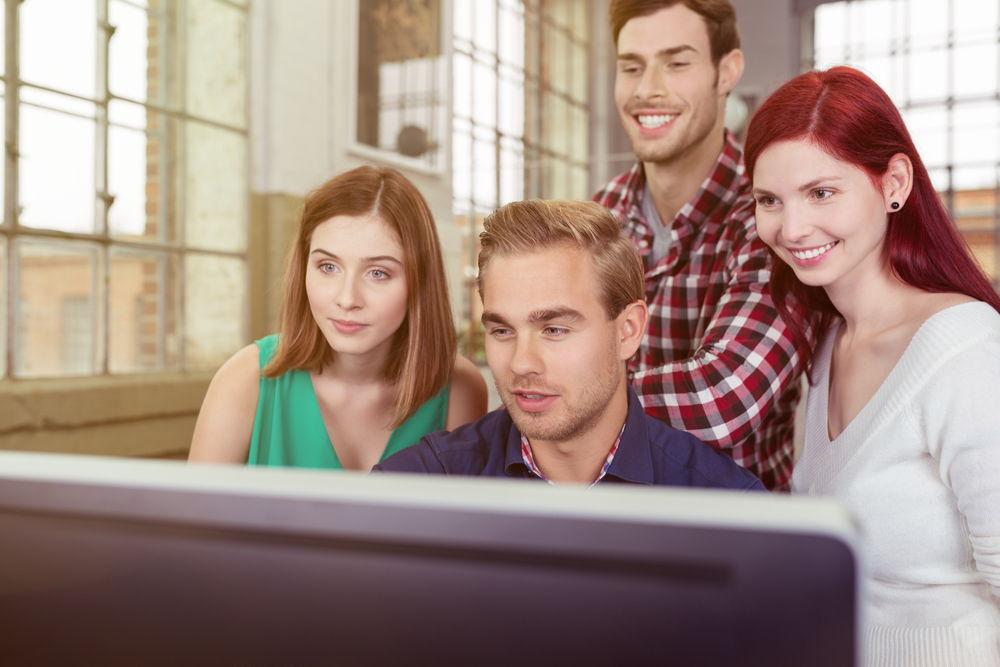 two men and two women in an office looking at a computer screen together